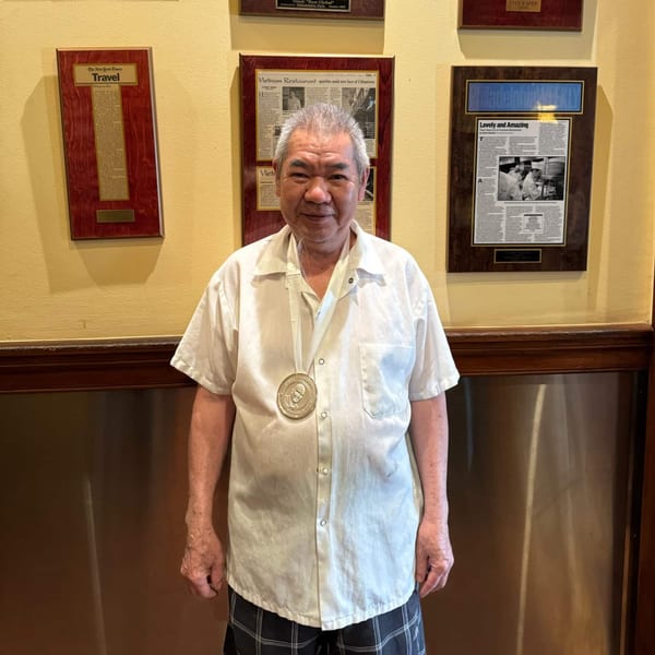 An older man in a white shirt and medal stands in front of framed newspaper clippings.