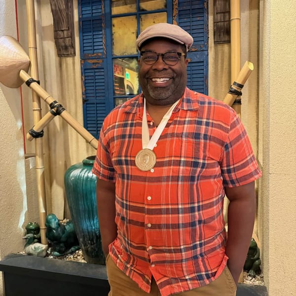 Smiling man wearing a medal, plaid shirt, and cap, standing in front of Vietnamese decor.