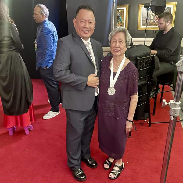 A man in a suit and an older woman in a purple dress with a medal pose on a red carpet.