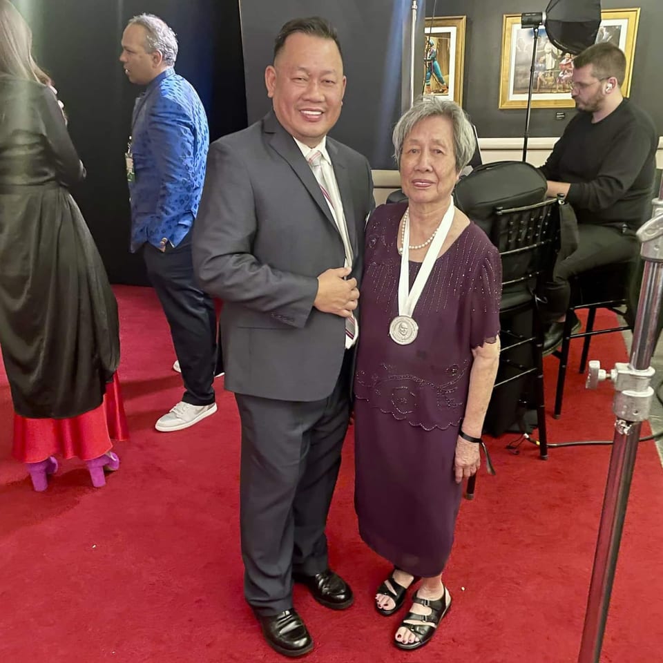 A man in a suit and an older woman in a purple dress with a medal pose on a red carpet.