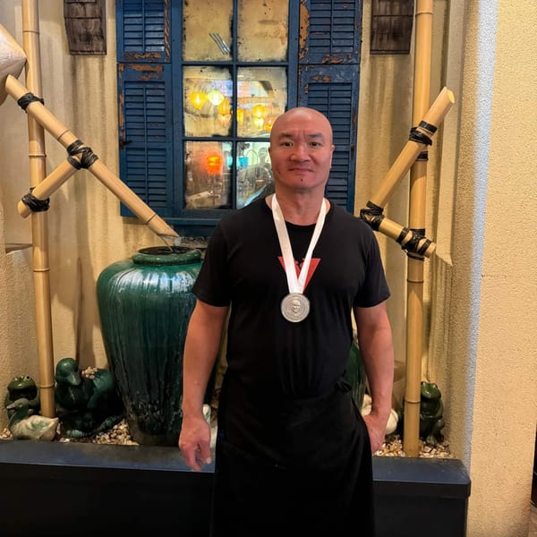A man wearing a James Beard Foundation medal stands in front of a blue window and bamboo decor.