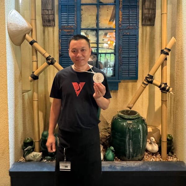 A man in a black apron and t-shirt with a red V logo holds a medal in a Vietnamese-themed setting.