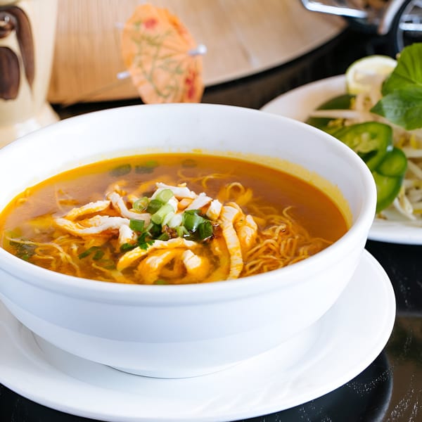 A bowl of Vietnamese chicken noodle soup with scallions, with a side of fresh vegetables.