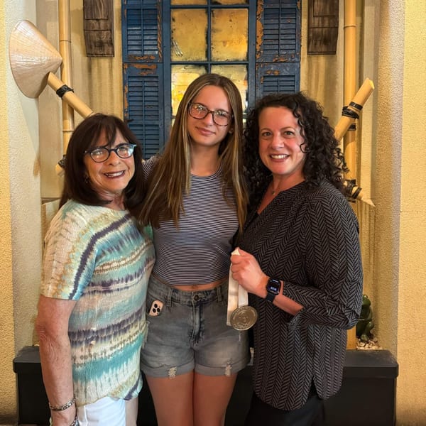 Three women smiling, standing in front of a yellow wall with Vietnamese decor.