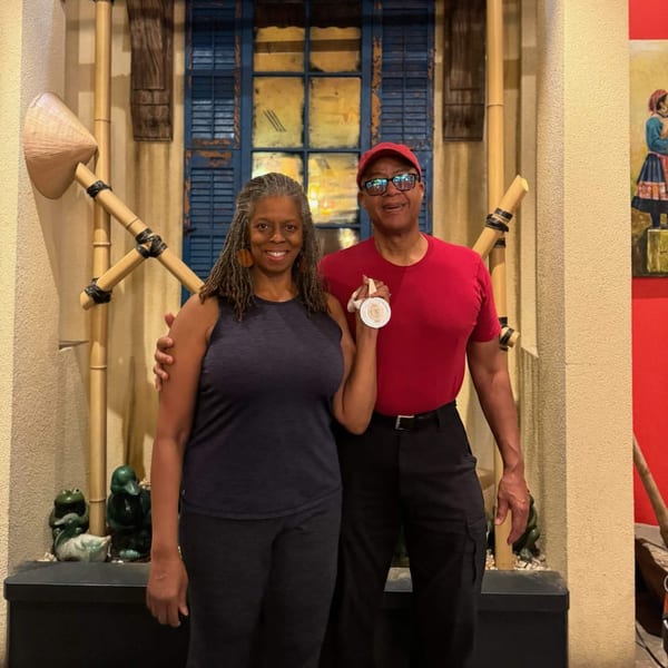 A smiling couple stands in front of a blue window with bamboo decor, holding a medal.