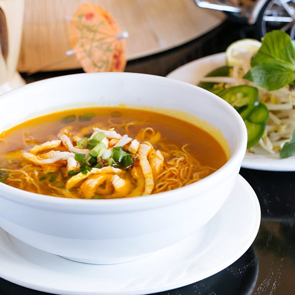 A bowl of Vietnamese chicken noodle soup with scallions, served with a side of fresh herbs.
