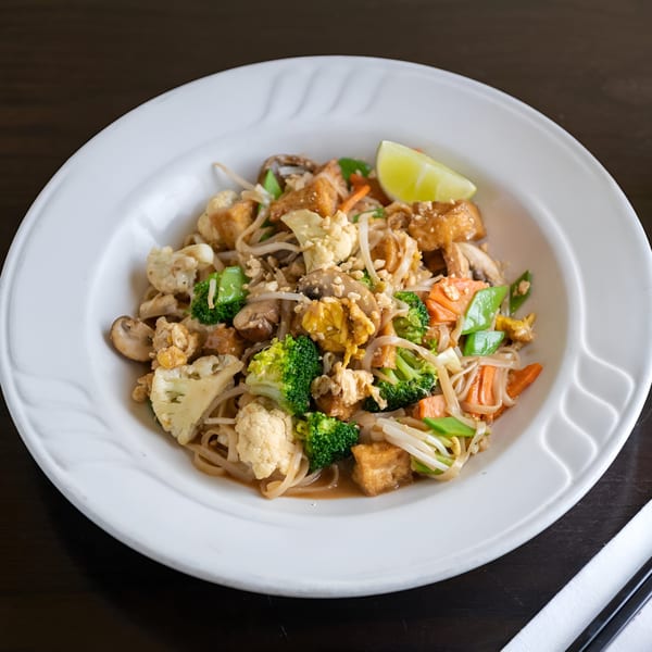 A Vietnamese noodle dish with tofu, broccoli, cauliflower, carrots, and mushrooms, garnished with a lime wedge.