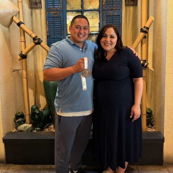 Man and woman smiling, man holding a medal, in front of a blue shuttered window and bamboo decor.