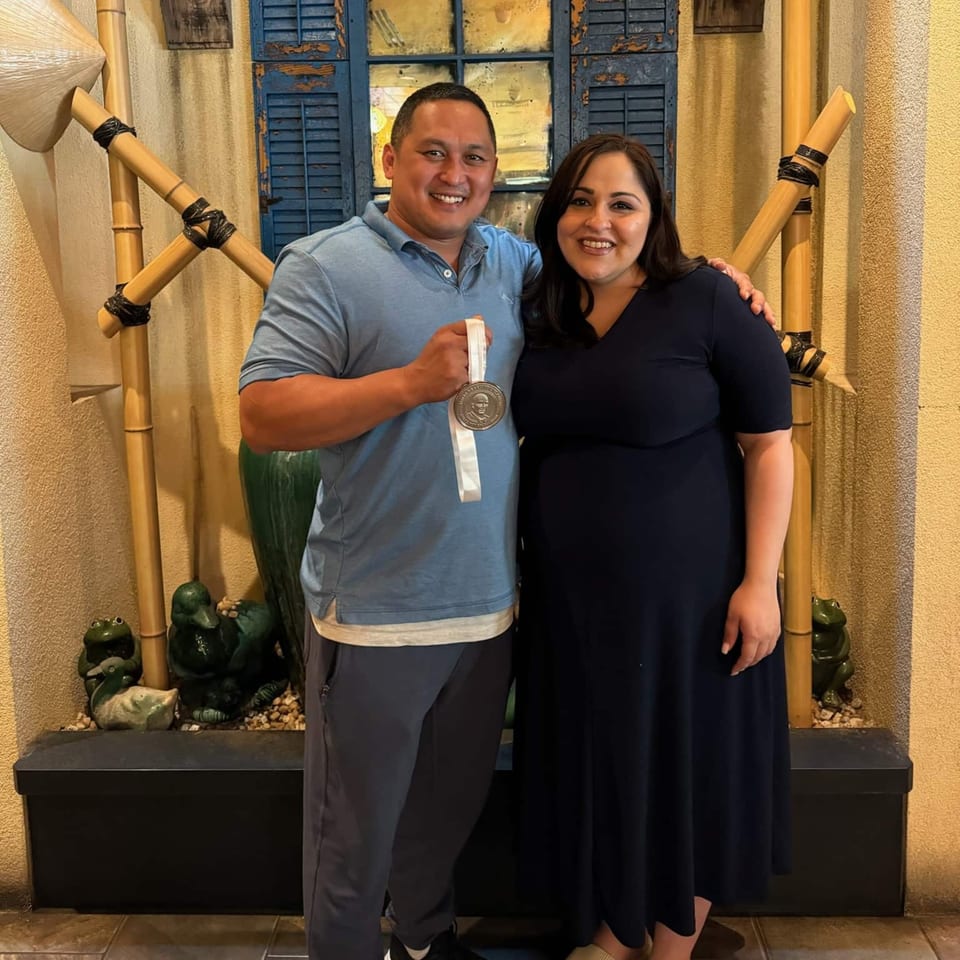 Man and woman smiling, man holding a medal, in front of a blue shuttered window and bamboo decor.