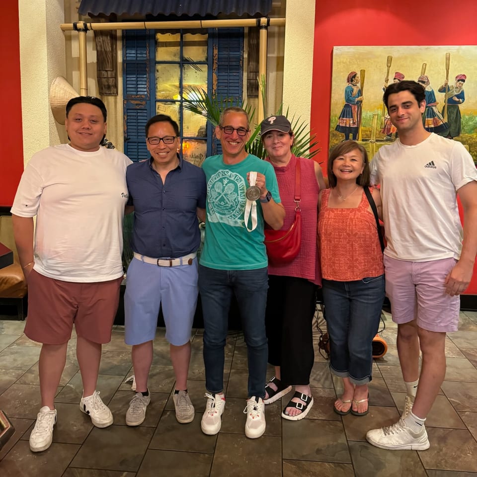 Six smiling people, including one holding a medal, stand in front of a Vietnamese-themed mural.